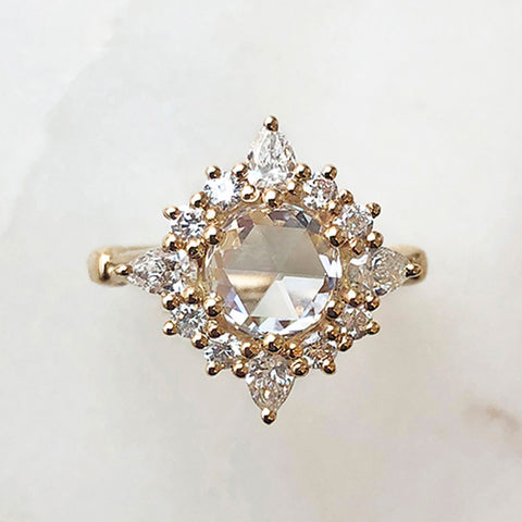 A cluster engagement ring with a rose cut white diamond center stone and round white diamond stones surrounding it set in yellow gold on a grey background.