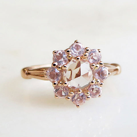 A cluster engagement ring with a rose cut morganite center stone and round pink morganite stones surrounding it set in rose gold on a grey background.