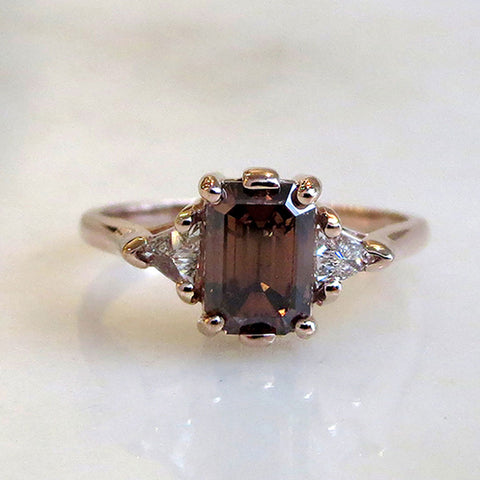 A three stone engagement ring with an emerald shape champagne diamond center stone with two white diamond trillion side stones set in yellow gold on a grey background.