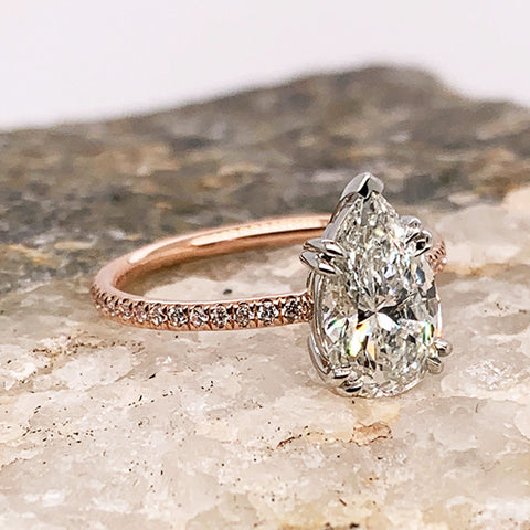 A solitaire pear white diamond ring with double white gold claw prongs with white diamond pave on the band set in rose gold on a marble stone.