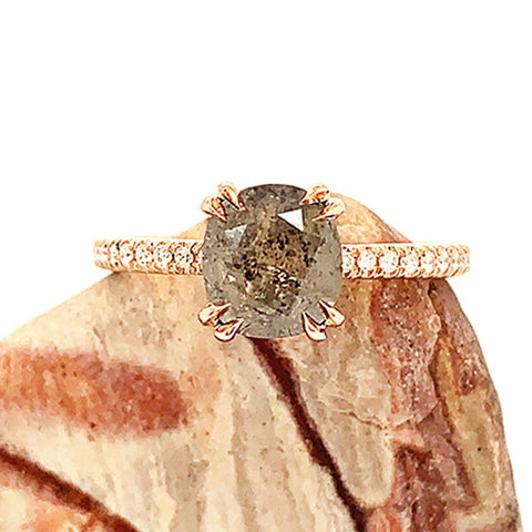 A solitaire cushion cut grey diamond ring with double claw prongs with white diamond pave on the band set in rose gold on a piece of wood.