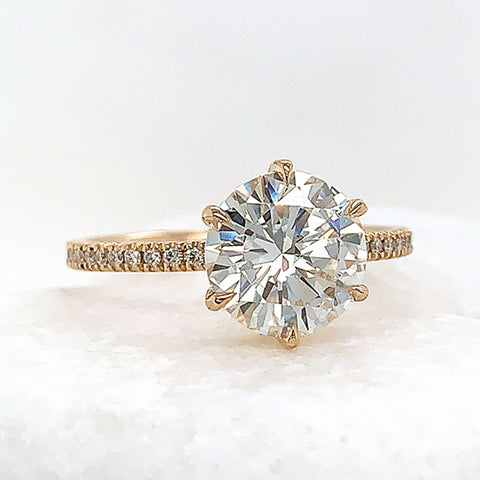 A solitaire round white diamond ring with claw prongs with white diamond pave on the band set in yellow gold on a white stone.