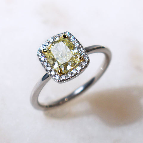 An engagement ring with an emerald shape yellow diamond center stone surrounded by a white diamond pave halo set in white gold on a grey background.