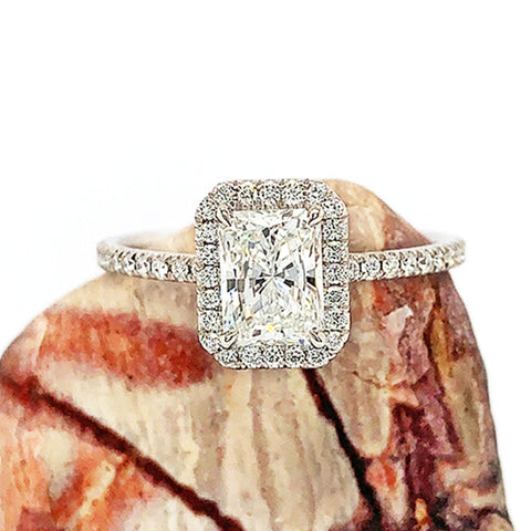 An engagement ring with an emerald shape white diamond center stone surrounded by a white diamond pave halo with white diamond pave around the band set in white gold on a marble stone.