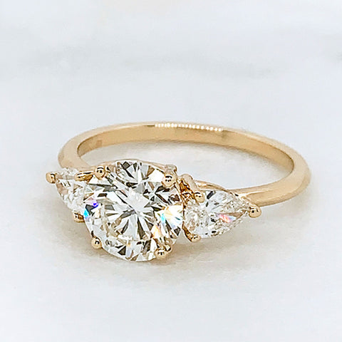 A three stone engagement ring with a round shape white diamond center stone with two white diamond pear shaped side stones set in yellow gold on a grey background.