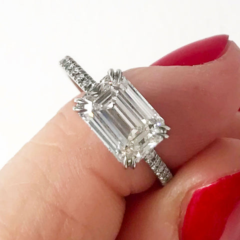A solitaire east/west emerald shape white diamond ring with double claw prongs with white diamond pave on the band set in white gold held by a light-complexion hand with red nails.