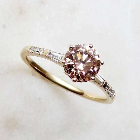 A solitaire ring with a round champagne diamond with tapered baguette accent stones and white diamond pave on either side set in yellow gold.