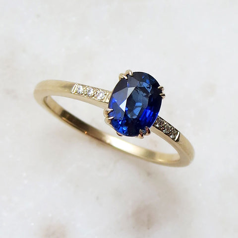 A solitaire ring with an oval blue sapphire stone with white diamond pave on either side set in yellow gold on a grey background.