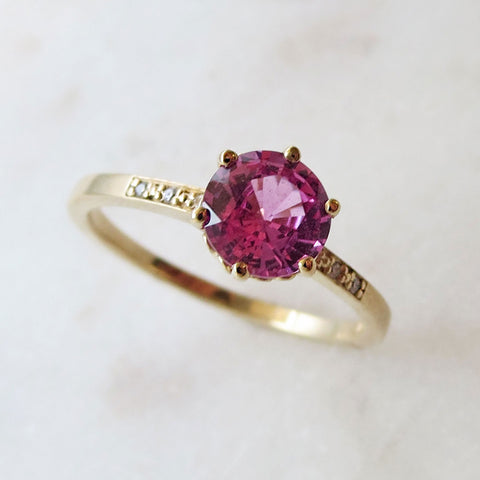 Angled view of a solitaire ring with a round pink sapphire stone with white diamond pave on either side set in yellow gold on a grey background.