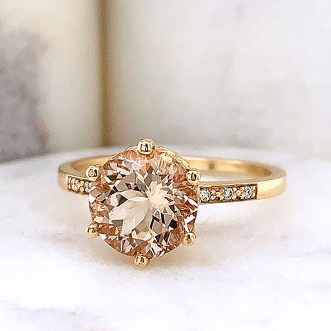 A solitaire ring with a round morganite stone with white diamond pave on either side set in yellow gold on a white stone.