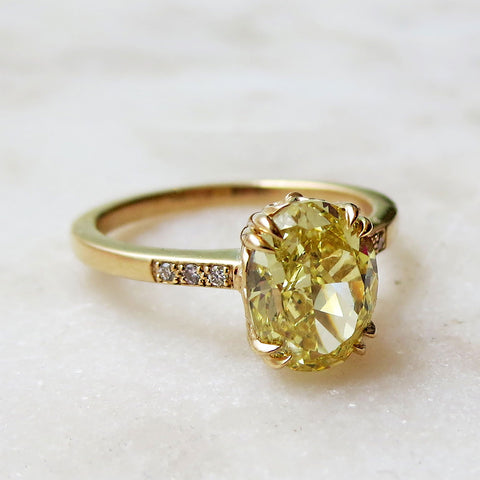 A solitaire ring with a cushion cut yellow diamond with white diamond pave on either side with double claw prongs set in yellow gold on a grey background.