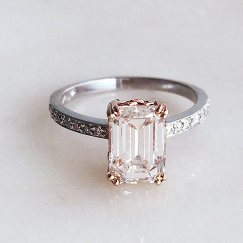 A solitaire ring with an emerald cut white diamond with white diamond pave on either side with double claw prongs in a rose gold basket set in white gold band on a grey background.