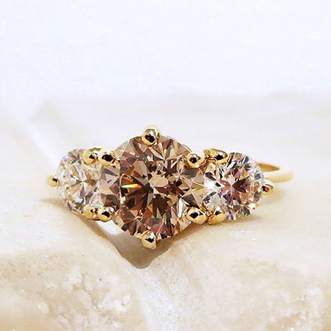 A three stone engagement ring with a round shape champagne diamond center stone with two white diamond round side stones set in yellow gold on a pink stone.
