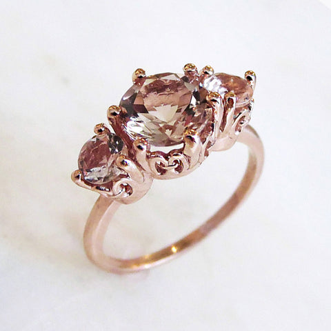 Angled view of a three stone engagement ring with a round shape pink morganite center stone with two pink morganite round side stones set in rose gold.
