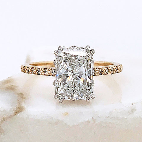 A solitaire emerald cut and shaped white diamond ring with double claw prongs set in white gold basket with white diamond pave on the band set in yellow gold on a grey stone.