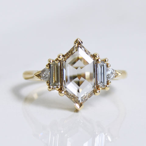 A ring with a hexagon shaped champange diamond center stone with one baguette white diamond and one white diamond trillion on either side set in yellow gold on a grey background.