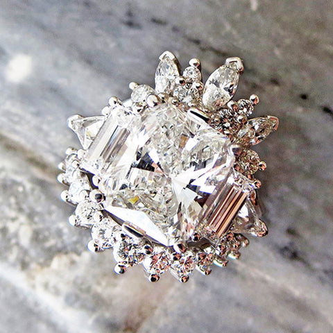 A ring with a brilliant cut and emerald shaped white diamond center stone with one baguette white diamond and one white diamond trillion on either side set in white gold stacked between two white diamond curve bands on a grey marble stone.