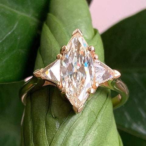 A three stone engagement ring with a marquise shape champagne diamond center stone with two white diamond trillion side stones set in yellow gold set in between greenery.