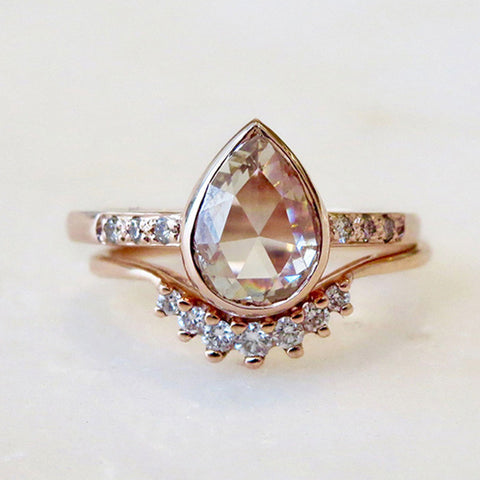 A solitaire ring with a pear rose cut champagne diamond with white diamond pave on either side set in rose gold above a curved band with 7 white diamonds in ascending and descending size set in rose gold on a grey background.