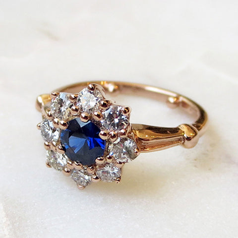 A cluster engagement ring with a rose cut blue sapphire center stone and round white diamond stones surrounding it set in yellow gold on a grey background.