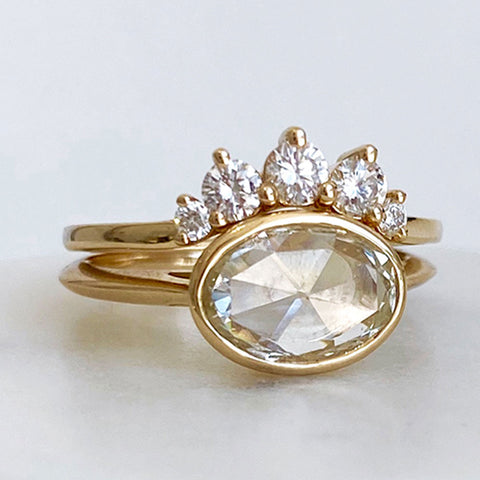An oval rose cut bezel set grey diamond set in yellow gold below a 5 white diamond curve band.