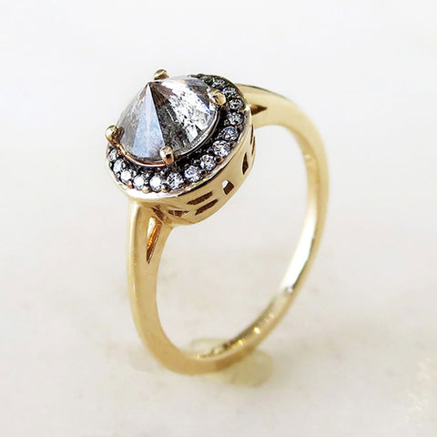 Angled view of an engagement ring with a round shape inverted grey diamond center stone surrounded by a white diamond pave halo with black rhodium set in yellow gold on a grey background.