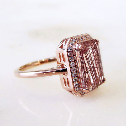 Angled view of an engagement ring with an emerald shape copper rutilated quartz center stone surrounded by a white diamond pave halo set in rose gold on a grey background.