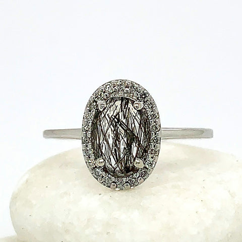 An engagement ring with an oval shape black rutilated quartz center stone surrounded by a white diamond pave halo set in white gold band on three white stones.