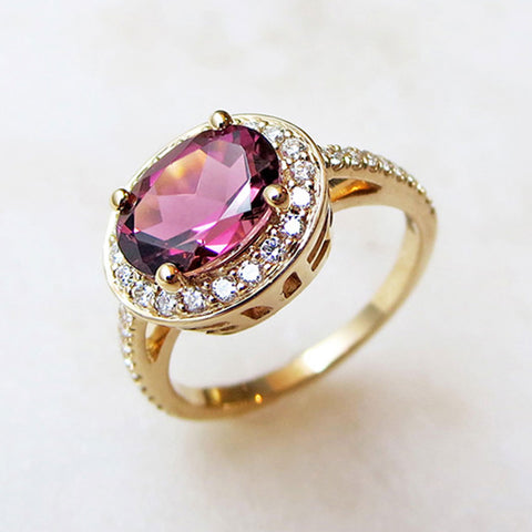 Angled view of an engagement ring with an oval shape pink tourmaline center stone surrounded by a white diamond pave halo set in yellow gold with white diamond pave around the band on a grey background.