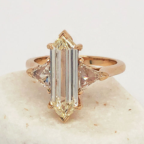 A three stone engagement ring with an hexagonal shape white diamond center stone with two white diamond trillion side stones set in yellow gold on a white rock.