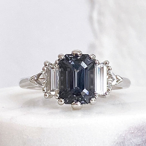 A ring with an emerald cut and shaped grey spinel center stone with one baguette white diamond and one white diamond trillion on either side set in white gold on a white marble stone.