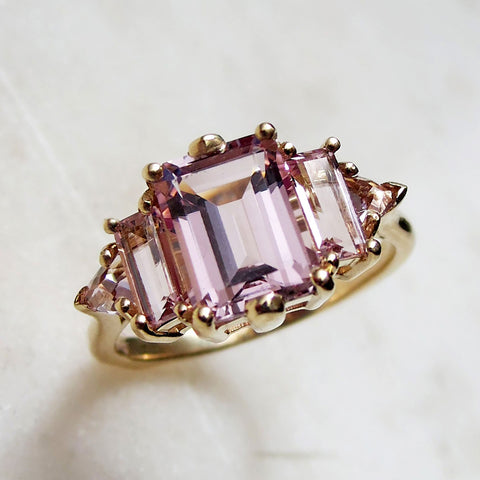 Angled view of a ring with an emerald cut and shaped pink morganite center stone with one baguette pink morganite and one pink morganite trillion on either side set in yellow gold on a grey background.