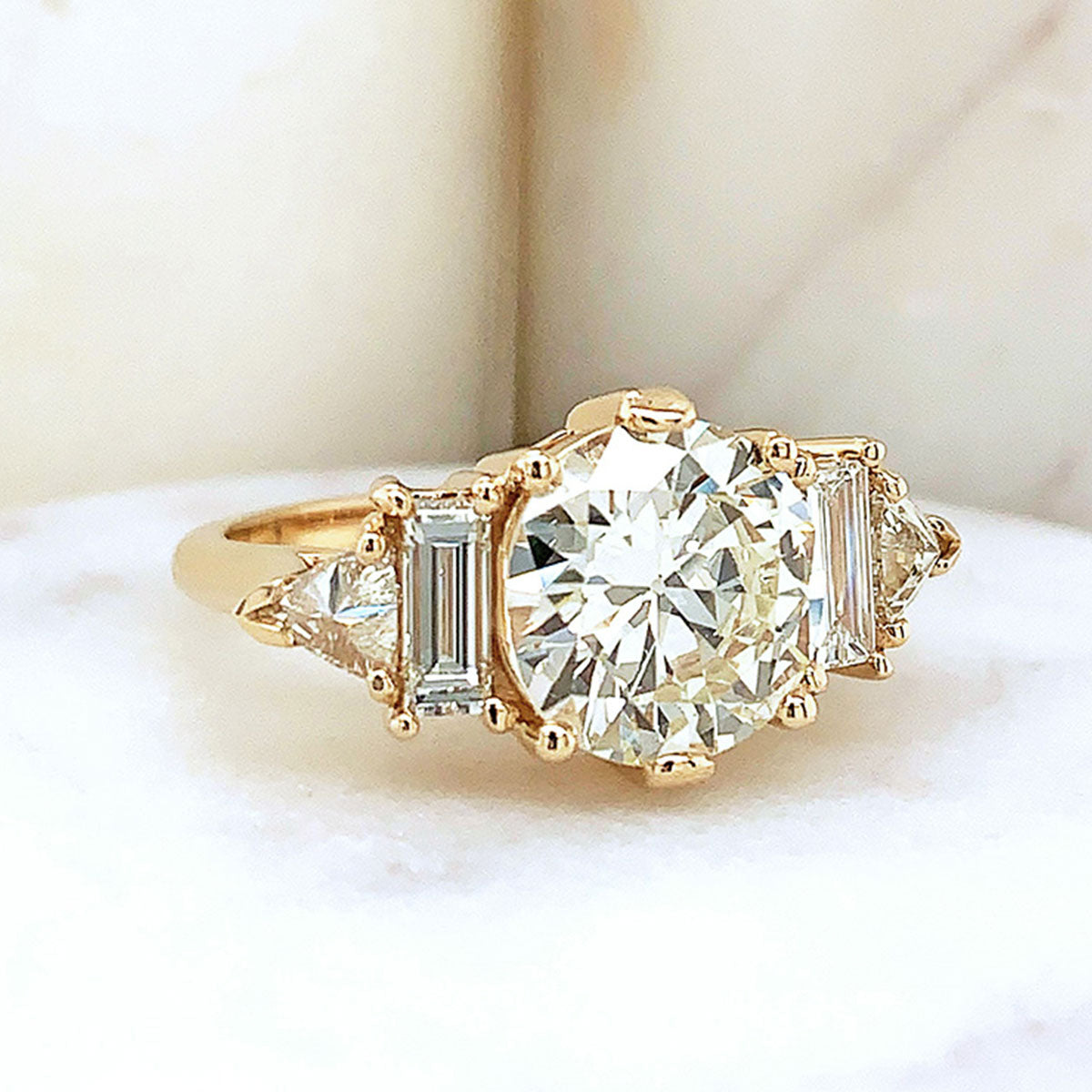 A ring with a round shaped white diamond center stone with one baguette white diamond and one white diamond trillion on either side set in yellow gold on a white stone.