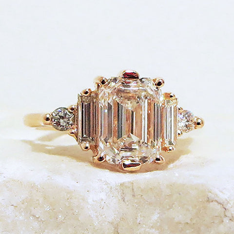 A ring with an emerald cut and shaped white diamond center stone with one baguette white diamond and one white diamond trillion on either side set in yellow gold on a grey stone.