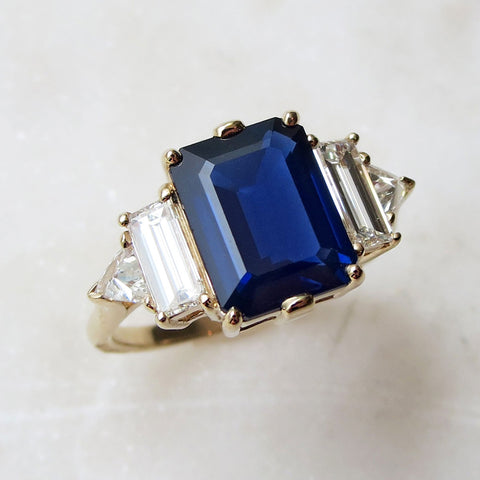Angled view of a ring with an emerald shaped blue sapphire center stone with one baguette white diamond and one white diamond trillion on either side set in yellow gold on a grey background.