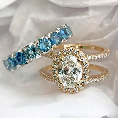 A double banded bezel set oval white diamond ring with a white diamond halo and white diamond pave around the band set in yellow gold with a blue gemstone eternity band set in white gold on a white fabric background.