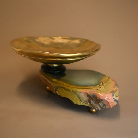 A golden bronze dish atop  piece of jasper with green, yellow, and pink striations on the gemstone on a beige background