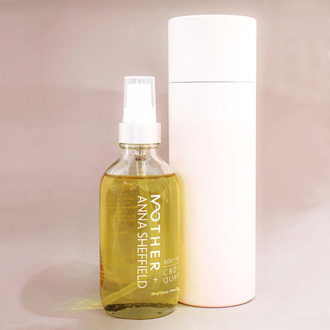 A bottle of CBD oil with "MOTHER + Anna Sheffield" written on it. Next to a cylinder for packaging and on a pink/grey background.
