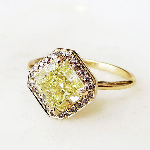 Angled view of an engagement ring with a square shape yellow diamond center stone surrounded by a champagne diamond pave halo set in yellow gold on a grey background.