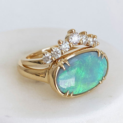 An oval bezel set opal with double claw prongs set in yellow gold with a 7 white diamond curve band stacked on top on a grey background.