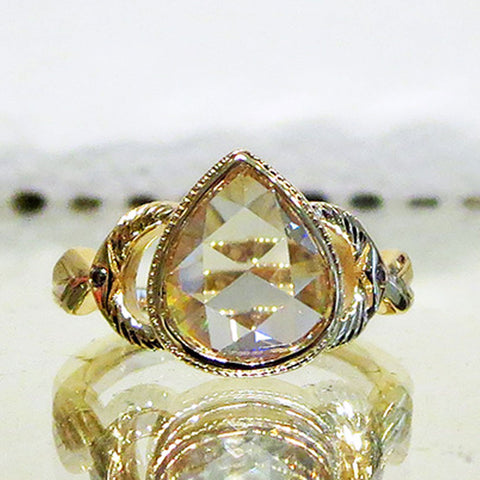 A pear rose cut white diamond with engraved gold accents on either side set in yellow gold on a mirror.