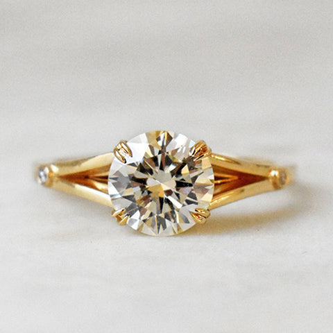 A ring with a round white diamond with double claw prong with triangle accent gold on either side set in yellow gold on a grey background.