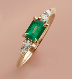 An angled view of a five stone ring with a green emerald center with two white diamonds on either side set in rose gold on a pink background.