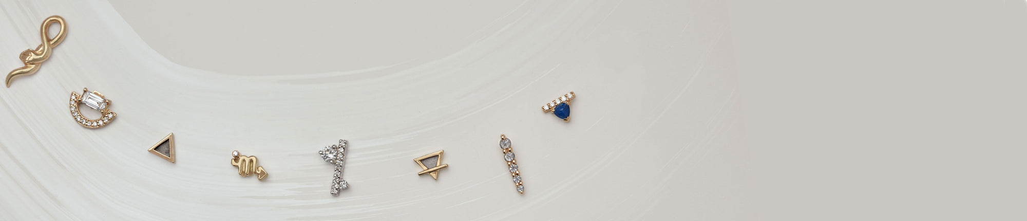 An array of 8 gold stud earrings in different shapes, stones, and colors on light-grey background with a brushstroke design
