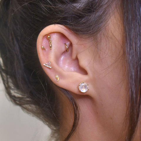 A woman's ear with multple gold stud earrings including a 7mm rainbow moonstone stud set in yellow gold.  2