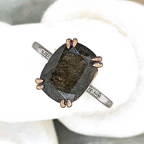 A solitaire ring with a cushion cut grey diamond with white diamond pave on either side set in rose gold basket and white gold band on three white stones.