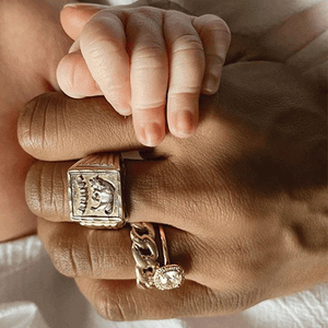 An adult hand with a mix of personal rings and an Anna Sheffield engagement ring hold the hand of a baby.