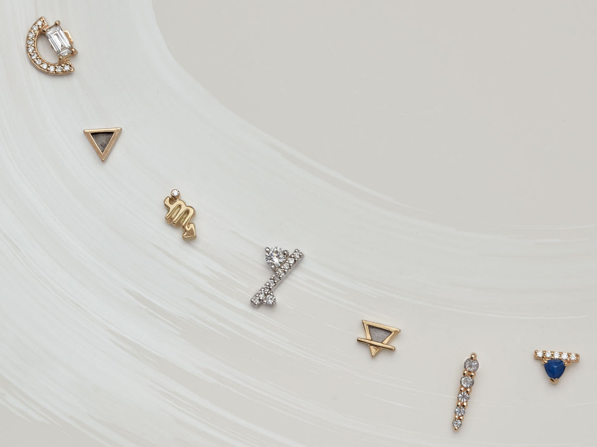 Zoomed in view of an array of 8 gold stud earrings in different shapes, stones, and colors on light-grey background with a brushstroke design