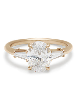A three stone engagement ring with an oval white diamond center stone and two white diamond tapered baguettes on either side set in yellow gold.