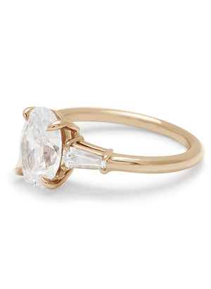Side view of a three stone engagement ring with an oval white diamond center stone and two white diamond tapered baguettes on either side set in yellow gold.